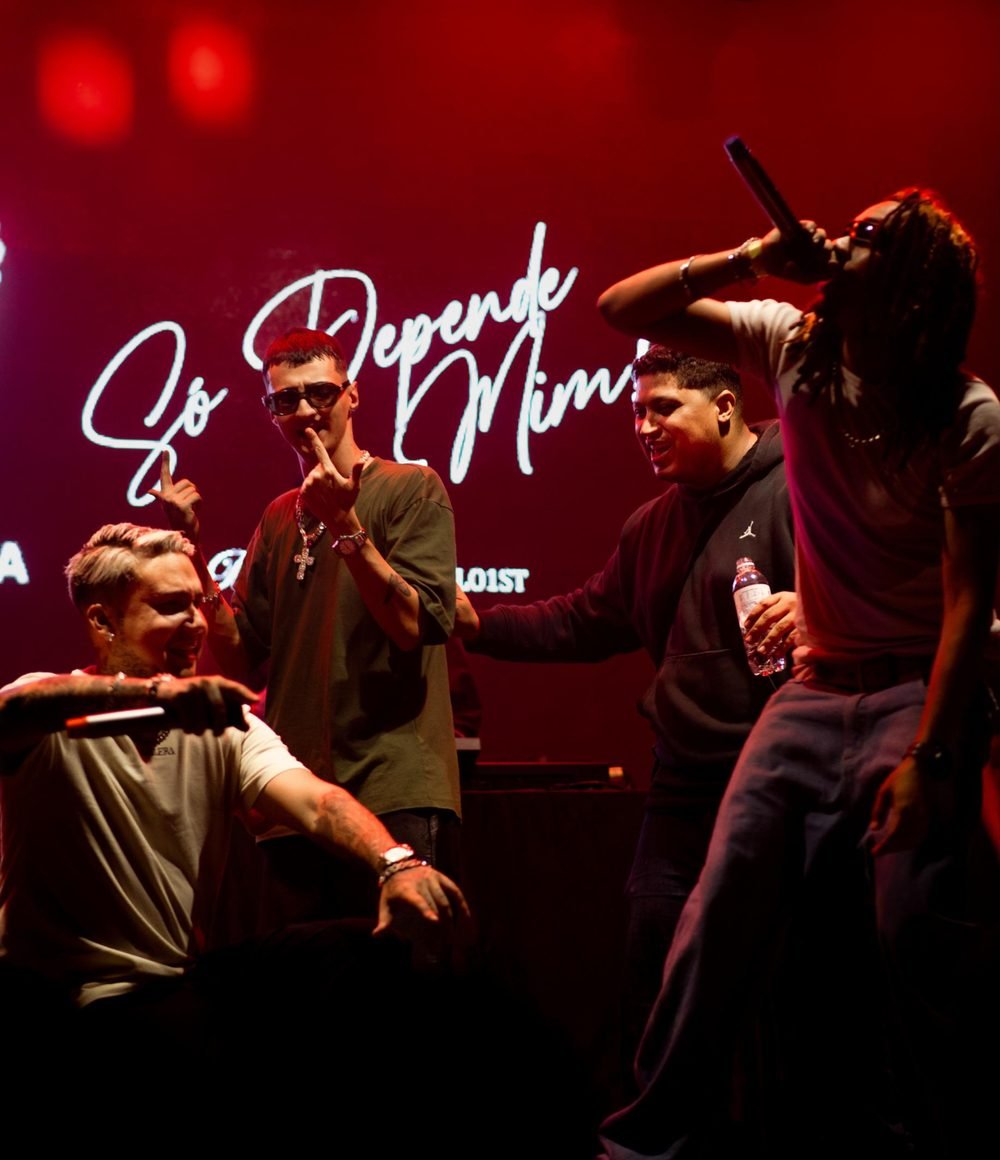 Quatro artistas no palco sob luz vermelha, telão com o nome Só Depende de Mim.